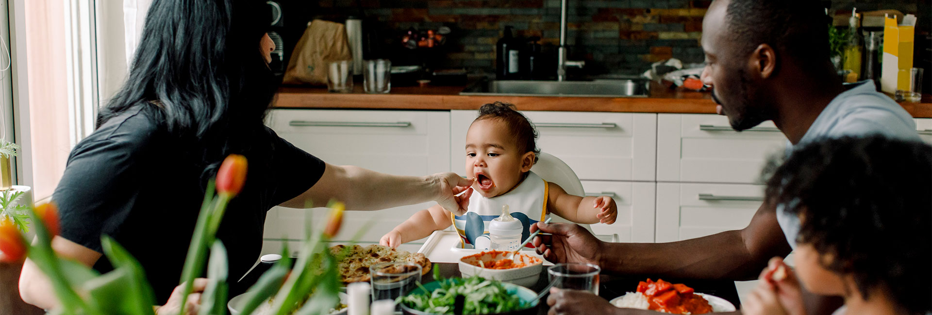 En kvinna matar en baby vid köksbordet med familjen runtom.